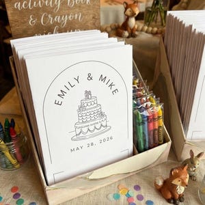 May include: A stack of white wedding activity books with a black line drawing of a three-tiered cake, the text EMILY & MIKE, and the date MAY 28, 2026. The books are in a cardboard display box alongside small clear bags of colorful crayons.