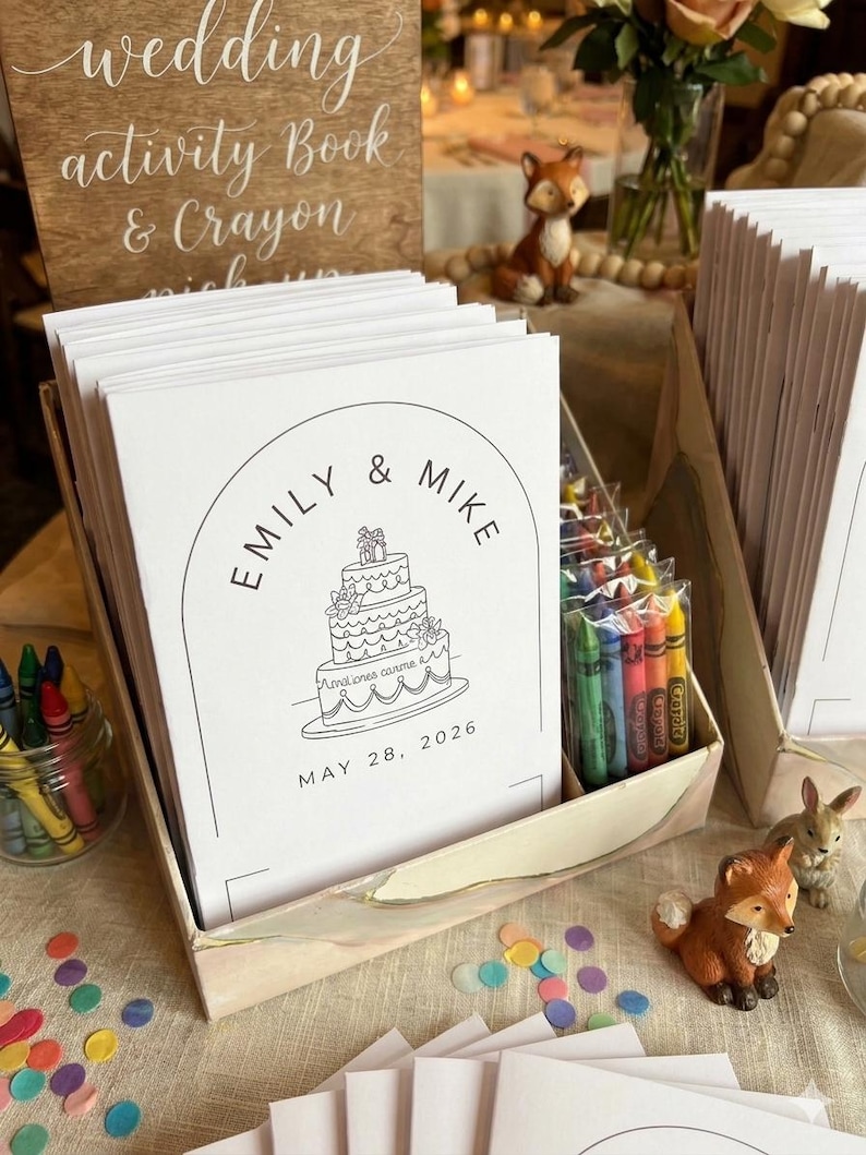 May include: A stack of white wedding activity books with a black line drawing of a three-tiered cake, the text EMILY & MIKE, and the date MAY 28, 2026. The books are in a cardboard display box alongside small clear bags of colorful crayons.