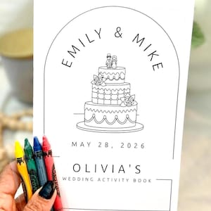 May include: A white rectangular wedding activity book with black line art featuring a three-tiered cake topped with a couple, arched framing, and the text EMILY & MIKE, MAY 28, 2026, and OLIVIA'S WEDDING ACTIVITY BOOK, accompanied by four crayons.