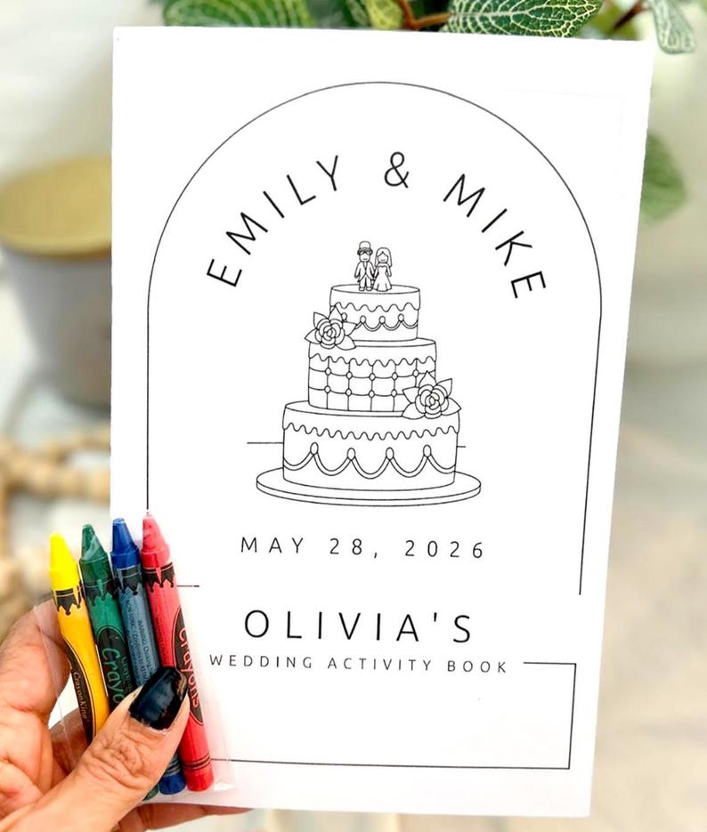 May include: A white rectangular wedding activity book with black line art featuring a three-tiered cake topped with a couple, arched framing, and the text EMILY & MIKE, MAY 28, 2026, and OLIVIA'S WEDDING ACTIVITY BOOK, accompanied by four crayons.