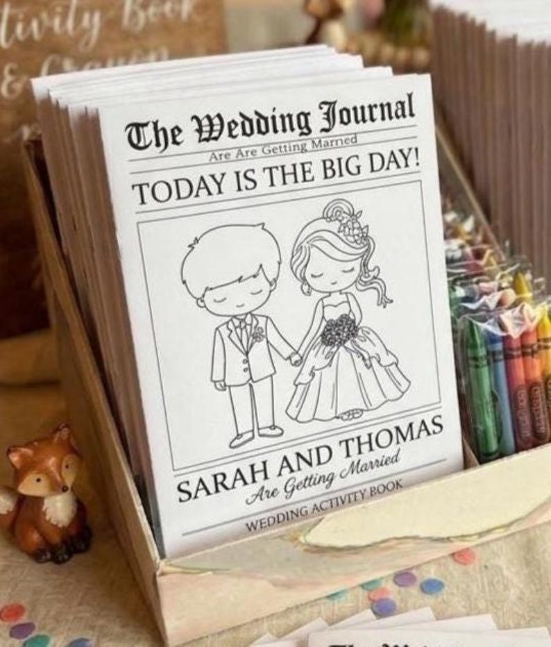 May include: A stack of white wedding activity books with black text and line art illustrations of a couple. The cover reads The Wedding Journal, TODAY IS THE BIG DAY!, and SARAH AND THOMAS Are Getting Married. A box of crayons sits beside the books.