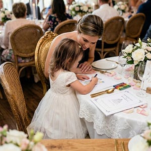 May include: A young child in a white dress with a lace bodice and tulle skirt sits at a round table, drawing on a paper with a black crayon. Beside the child, a wedding program reads Samantha & Michael, accompanied by gold cutlery and crayons.