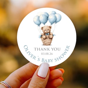 May include: A round sticker with a watercolor illustration of a teddy bear in a wicker basket, held aloft by light blue balloons. The text reads "THANK YOU" and "OLIVER'S BABY SHOWER".