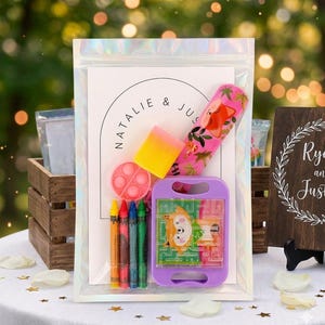 May include: A clear iridescent pouch containing a white card with the text NATALIE & JUSTIN, a pink fox-patterned ruler, a yellow and pink rectangular eraser, a small pink circular fidget toy, four crayons, and a purple handheld maze game.