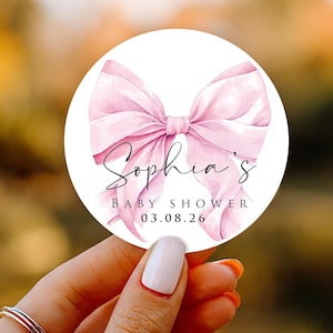 May include: A round sticker with a watercolor-style pink bow design. The sticker has the text "Sophia's BABY SHOWER 03.08.26" in a script font. The sticker is held by a person with a white manicure.