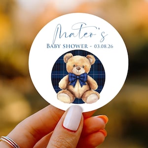 May include: A round sticker with the text "Mateo's BABY SHOWER - 03.08.26". The sticker features a teddy bear with a blue bow tie, set against a blue plaid background. The bear is light brown, and the text is in a cursive font.