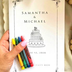 May include: A white activity book with black text reading SAMANTHA & MICHAEL and JUNE 12, 2026, featuring a line drawing of a three-tiered wedding cake with a topper. A small clear plastic sleeve contains four crayons in yellow, green, blue, and red.