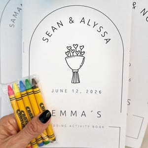 May include: A white wedding activity book with black text and a line drawing of a heart bouquet. The cover features the names Sean & Alyssa, the date June 12, 2026, and the text Emma's Wedding Activity Book. A small clear pack of five crayons is included.