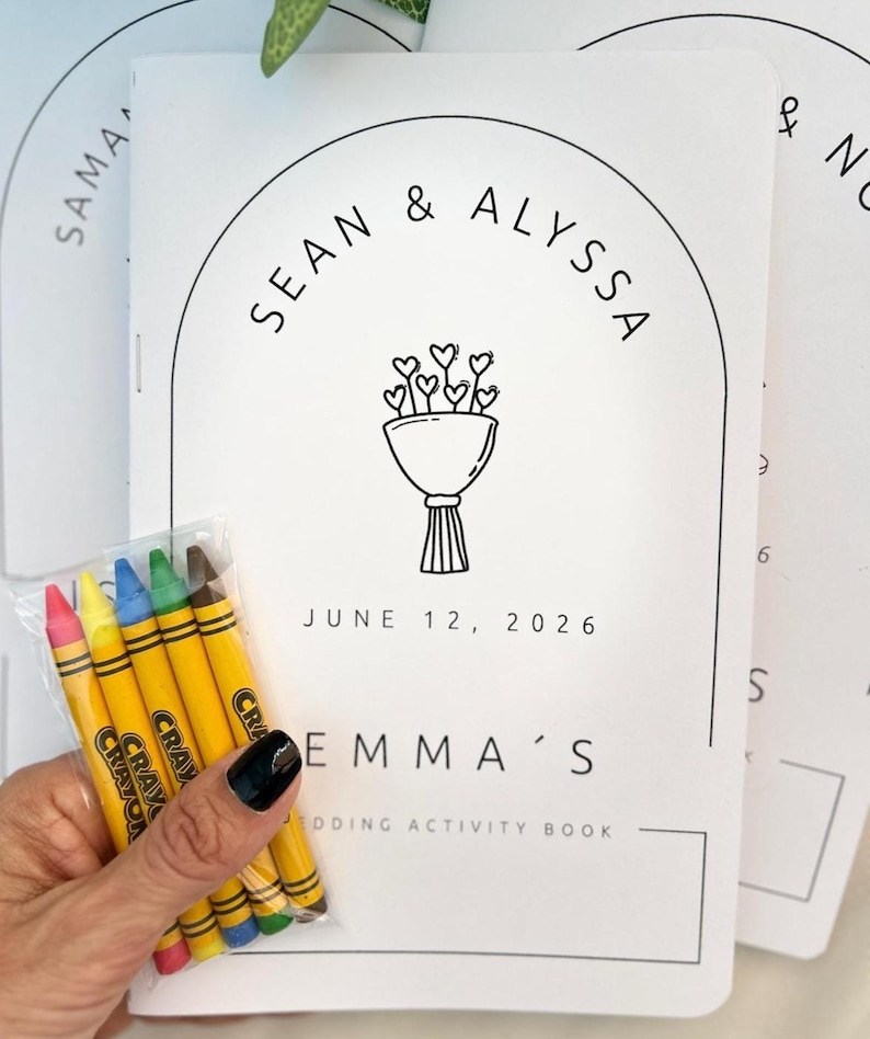 May include: A white wedding activity book with black text and a line drawing of a heart bouquet. The cover features the names Sean & Alyssa, the date June 12, 2026, and the text Emma's Wedding Activity Book. A small clear pack of five crayons is included.