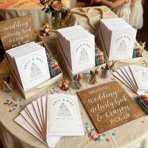 May include: Multiple stacks of white wedding activity books with a black line drawing of a tiered cake and the text Emily & Mike May 28, 2026. The books are displayed with wooden signs reading wedding activity book & crayon pick-up and jars of crayons.