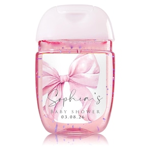 May include: Pink hand sanitizer bottle with a white cap. The bottle is decorated with a watercolor bow and the text "Sophia's BABY SHOWER 03.08.26". The bottle has a pink liquid with purple glitter.