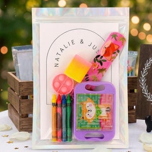 May include: A clear iridescent pouch containing a white card with the text Natalie & Jus, a pink fox-patterned tube, a yellow and pink eraser, a small pink circular fidget toy, four crayons in yellow, red, blue, and green, and a purple maze game.