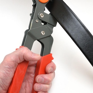 May include: A gray and orange belt hole punch tool is used on a black leather belt. The tool has orange handles and an orange circular piece. The belt is held in place by the tool.
