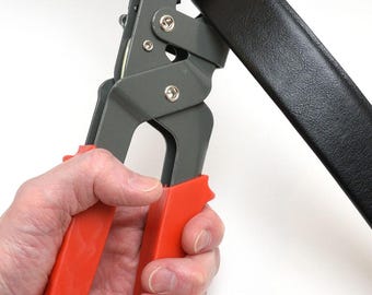 May include: A gray and orange belt hole punch tool is used on a black leather belt. The tool has orange handles and an orange circular piece. The belt is held in place by the tool.