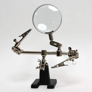 May include: A silver-colored helping hands tool with a magnifying glass and two alligator clips. The tool is mounted on a black base and has adjustable arms for holding small objects during soldering or other detailed work.