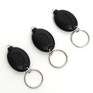May include: Three black LED keychain flashlights with silver key rings. Each flashlight has an oval shape with a button and a clear, rounded light at the top. The flashlights are arranged diagonally against a white background.