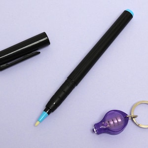 May include: A black pen with a light blue tip and cap, next to a black pen cap and a purple lightbulb keychain with a gold ring. The pen has a sleek design, and the keychain has a small lightbulb inside.