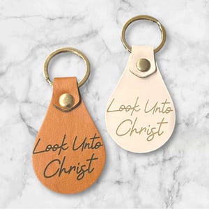 May include: Two leather keyrings with gold key rings. Both keychains have a teardrop shape and are embossed with the text "Look Unto Christ". One keychain is brown and the other is white.