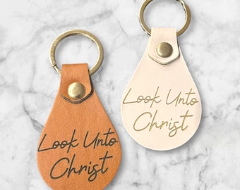 Puede incluir: Dos llaveros de cuero con anillas doradas. Ambos llaveros tienen forma de lágrima y están grabados con el texto "Look Unto Christ". Un llavero es marrón y el otro es blanco.