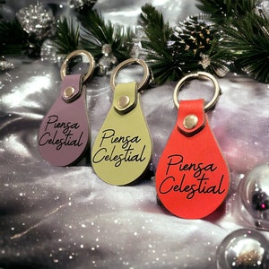 May include: Three leather keychains with silver key rings. Each keychain is a teardrop shape with a different color: purple, green, and red. The text "Piensa Celestial" is engraved on each keychain.