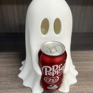 May include: A white 3D-printed ghost figure holding a red Dr. Pepper can. The ghost has two oval eye cutouts. The Dr. Pepper can has white lettering and the Jurassic World logo. The ghost is a fun, decorative item.