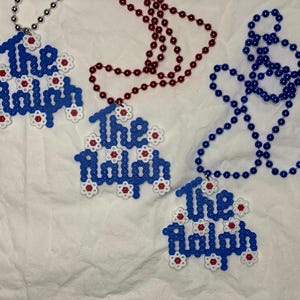 May include: Three beaded necklaces with pendants that read "The Ralph". The pendants are blue with white and red accents. One necklace is silver, one is red, and one is blue.