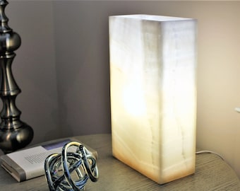 May include: A white alabaster table lamp with a rectangular base. The lamp is lit and casting a warm glow.