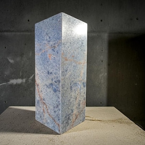 May include: A blue marble lamp with red veining, standing on a concrete surface. The rectangular lamp has a polished finish and a cord extending from the base. The background is a dark gray concrete wall.
