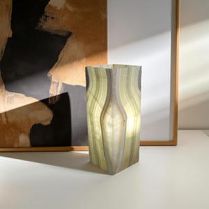 May include: A rectangular, translucent onyx lamp with a soft, glowing interior. The lamp features vertical bands of green, white, and brown. The lamp is placed on a white surface, with abstract art and a framed picture in the background.