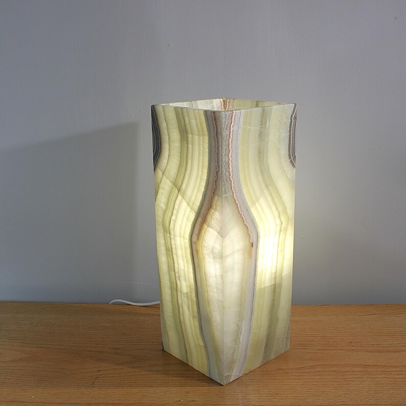 Puede incluir: L&aacute;mpara rectangular de &oacute;nix iluminada, con un patr&oacute;n verde y blanco. La l&aacute;mpara tiene una base cuadrada y una superficie texturizada. Se ve un cable de alimentaci&oacute;n. La luz brilla a trav&eacute;s de la piedra, creando un brillo c&aacute;lido.