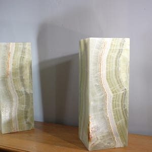 Puede incluir: Dos esculturas rectangulares de &oacute;nix con un patr&oacute;n de capas verde claro y blanco. Los bloques de piedra est&aacute;n de pie sobre una superficie de madera, con una pared gris al fondo. Las esculturas tienen una est&eacute;tica natural y org&aacute;nica.