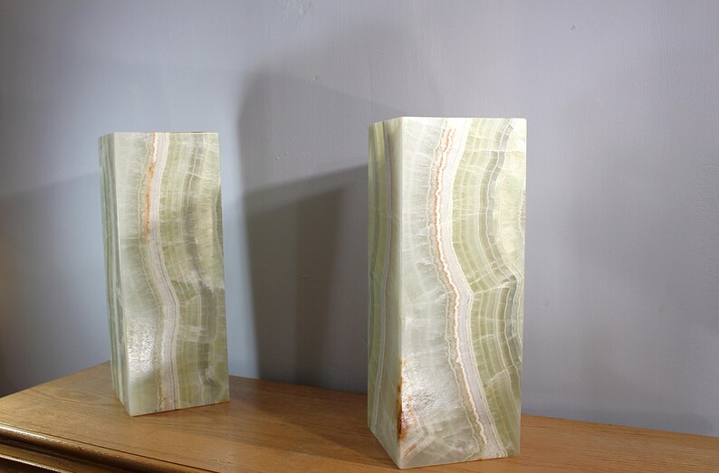 Puede incluir: Dos esculturas rectangulares de &oacute;nix con un patr&oacute;n de capas verde claro y blanco. Los bloques de piedra est&aacute;n de pie sobre una superficie de madera, con una pared gris al fondo. Las esculturas tienen una est&eacute;tica natural y org&aacute;nica.