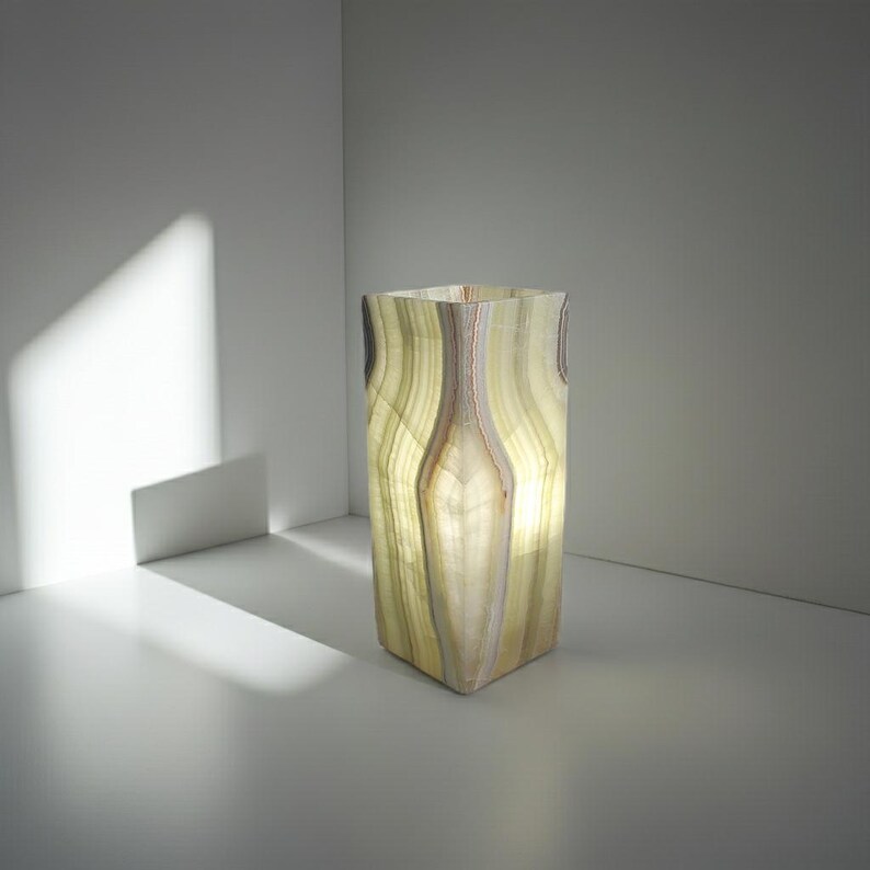 Puede incluir: Un jarr&oacute;n rectangular de &oacute;nix iluminado con un patr&oacute;n de rayas verdes claras y marrones. La piedra transl&uacute;cida permite el paso de la luz, creando un brillo suave. El jarr&oacute;n est&aacute; colocado en un entorno minimalista.