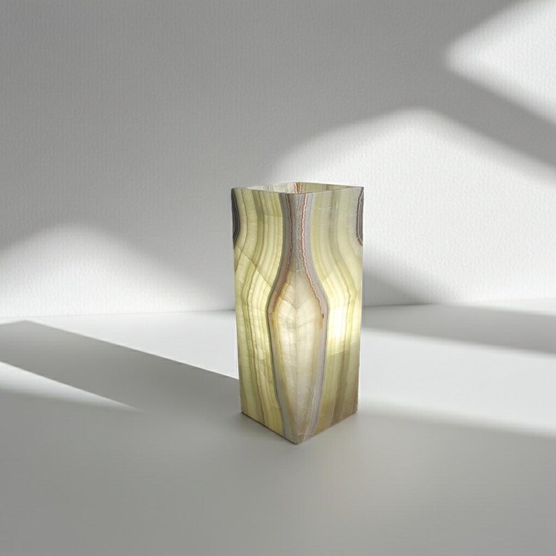 Puede incluir: Una l&aacute;mpara rectangular de &oacute;nix transl&uacute;cido con un interior suave y brillante. La l&aacute;mpara presenta bandas verticales de verde claro, crema y marr&oacute;n, creando un efecto natural y estratificado. La l&aacute;mpara mide aproximadamente 15 cm de alto.