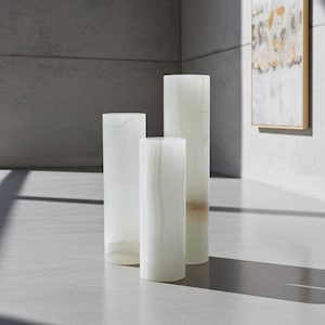 May include: Three cylindrical decorative vases in varying heights. The vases are made of a translucent, off-white material with subtle veining. The tallest vase has a slight tan hue at the base. The vases are arranged on a light gray surface.