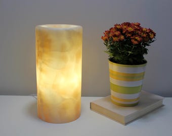 May include: A white alabaster lamp with a cylindrical shape and a warm glow. The lamp is sitting on a white table next to a book and a potted plant with orange flowers.