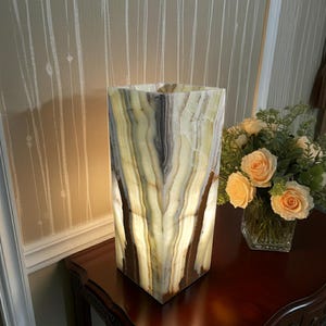 May include: A rectangular onyx lamp with a warm glow. The lamp features vertical bands of cream, brown, and gray. The lamp is on a dark wood table next to a vase of peach roses.
