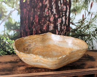 May include: A large, oval-shaped, natural stone bowl with a light brown, beige, and white marbled pattern. The bowl is sitting on a wooden surface.