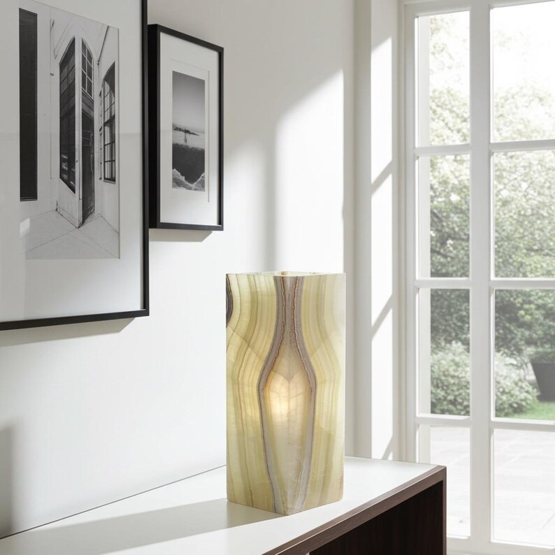 Puede incluir: L&aacute;mpara rectangular de &oacute;nix iluminada, con un patr&oacute;n natural en capas de tonos crema, beige y verde. La l&aacute;mpara est&aacute; colocada sobre una superficie blanca, con obras de arte en blanco y negro enmarcadas en el fondo.