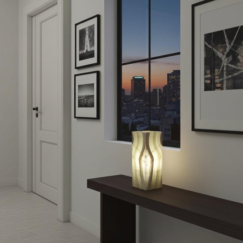 Puede incluir: Una l&aacute;mpara decorativa iluminada con un patr&oacute;n de rayas beige y marr&oacute;n. La l&aacute;mpara rectangular est&aacute; sobre una mesa marr&oacute;n oscuro. El fondo incluye obras de arte enmarcadas, una ventana y una puerta.
