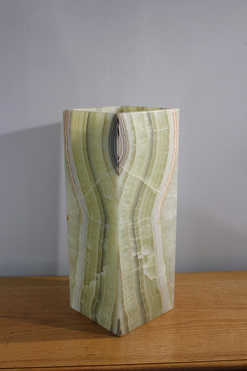 Puede incluir: Un jarr&oacute;n alto y rectangular de &oacute;nix verde con un patr&oacute;n &uacute;nico en capas. El jarr&oacute;n tiene una parte superior e inferior cuadradas, y la piedra natural muestra tonos de verde, blanco y marr&oacute;n. El jarr&oacute;n mide unos 30 cm de alto.