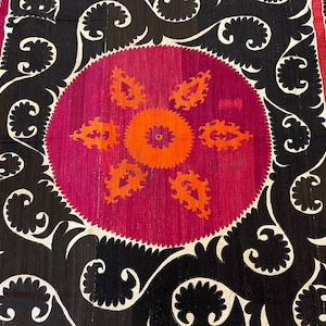 May include: A close-up of a textile with a central circular design in magenta and orange, surrounded by black and white ornate patterns. The textile features a repeating floral border in red and orange.