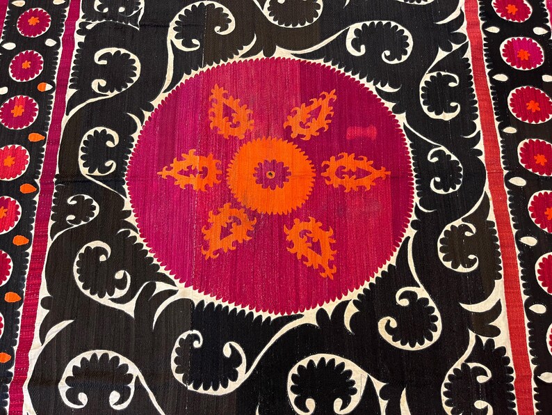 May include: A close-up of a textile with a central circular design in magenta and orange, surrounded by black and white ornate patterns. The textile features a repeating floral border in red and orange.