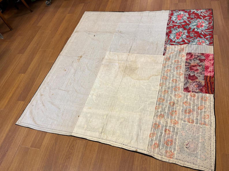 May include: A patchwork quilt with various fabric patterns. The quilt features sections of white and gray striped fabric, floral prints in red and pink, and a cream-colored central panel. The quilt is laid out on a wooden floor.