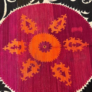 May include: Close-up of a textile with a central circular design. The design features a vibrant orange and purple floral pattern on a magenta background. The border is black with white, curved accents. The textile has a textured appearance.