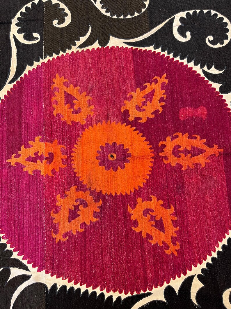 May include: Close-up of a textile with a central circular design. The design features a vibrant orange and purple floral pattern on a magenta background. The border is black with white, curved accents. The textile has a textured appearance.