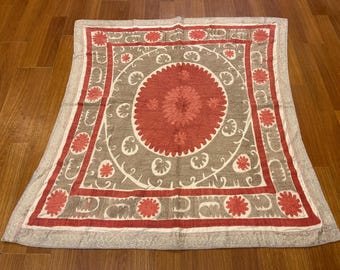 May include: A rectangular textile wall hanging featuring a large central coral-red medallion surrounded by a taupe field with white swirling vine patterns. The border is defined by coral-red bands with repeating floral motifs.