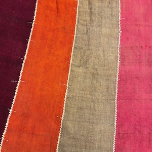 May include: A close-up of a textile with vertical stripes in shades of burgundy, orange, tan, and pink. The stripes are separated by white zigzag stitching, creating a textured and colorful pattern. The fabric appears to be woven.