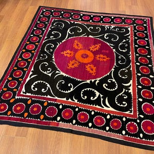 May include: A square, decorative textile with a central circular design in shades of fuchsia and orange, surrounded by black and white swirling patterns. The border features repeating floral motifs in red and orange.