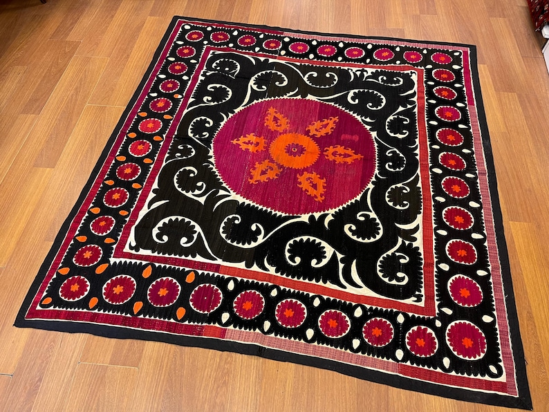 May include: A square, decorative textile with a central circular design in shades of fuchsia and orange, surrounded by black and white swirling patterns. The border features repeating floral motifs in red and orange.